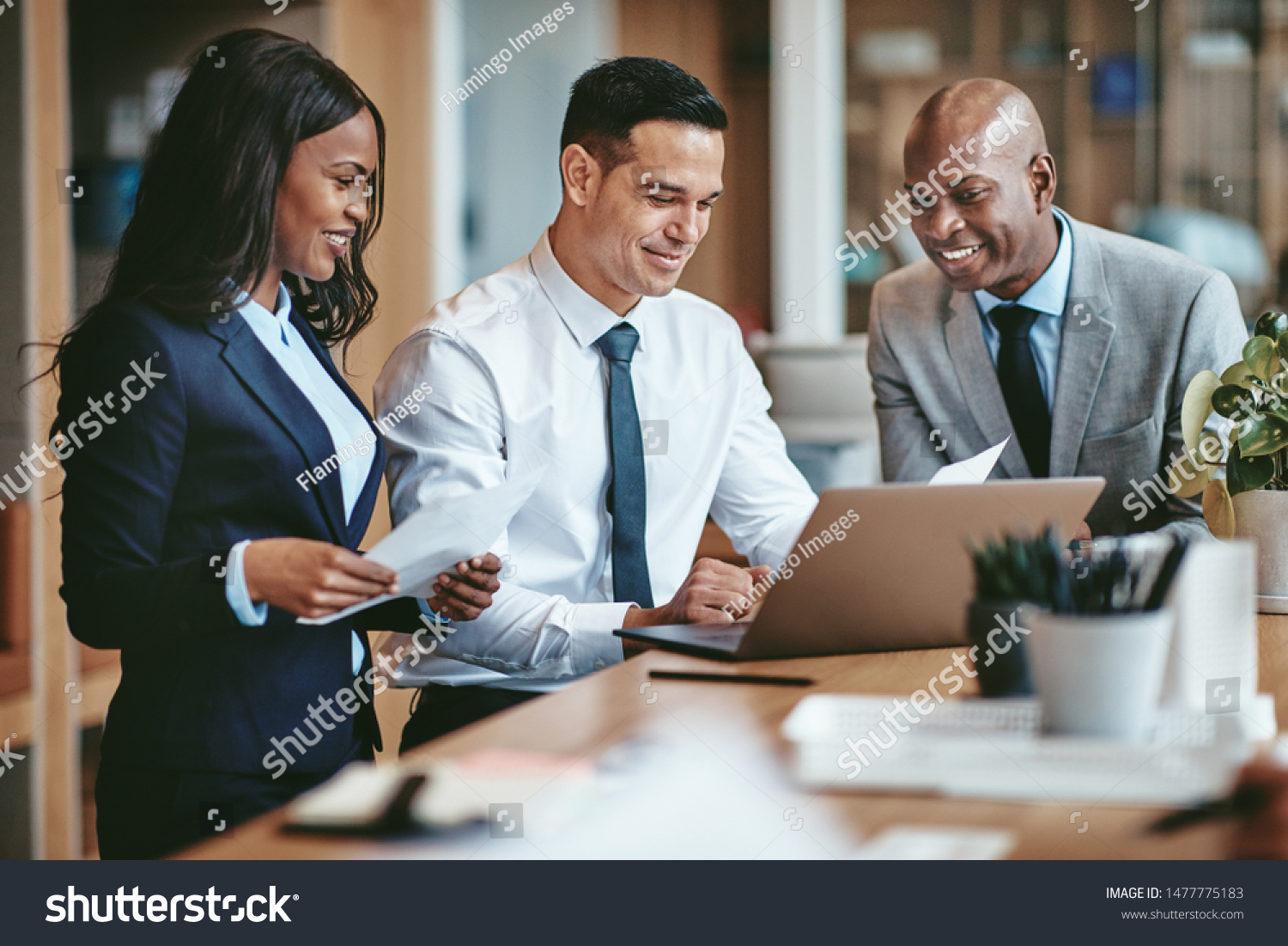 Smiling group of diverse businesspeople going over paperwork together and working on a laptop at a table in an office #1477775183