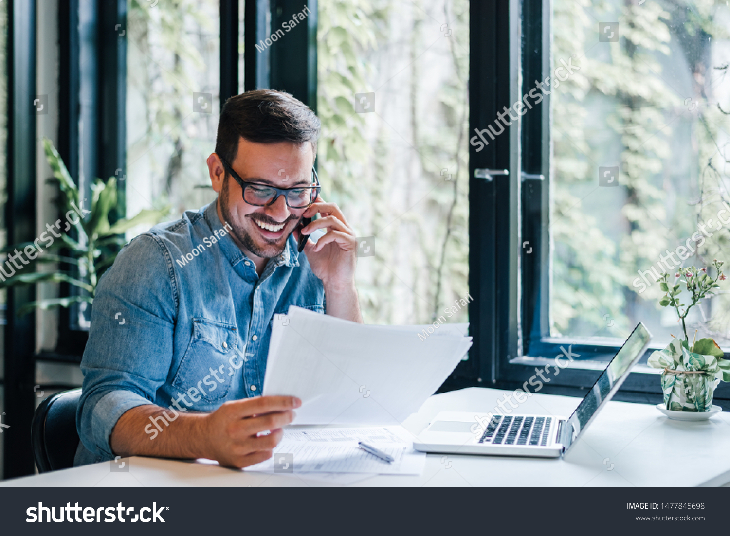 Portrait of young smiling cheerful entrepreneur in casual office making phone call while working with charts and graphs looking at paper documents #1477845698