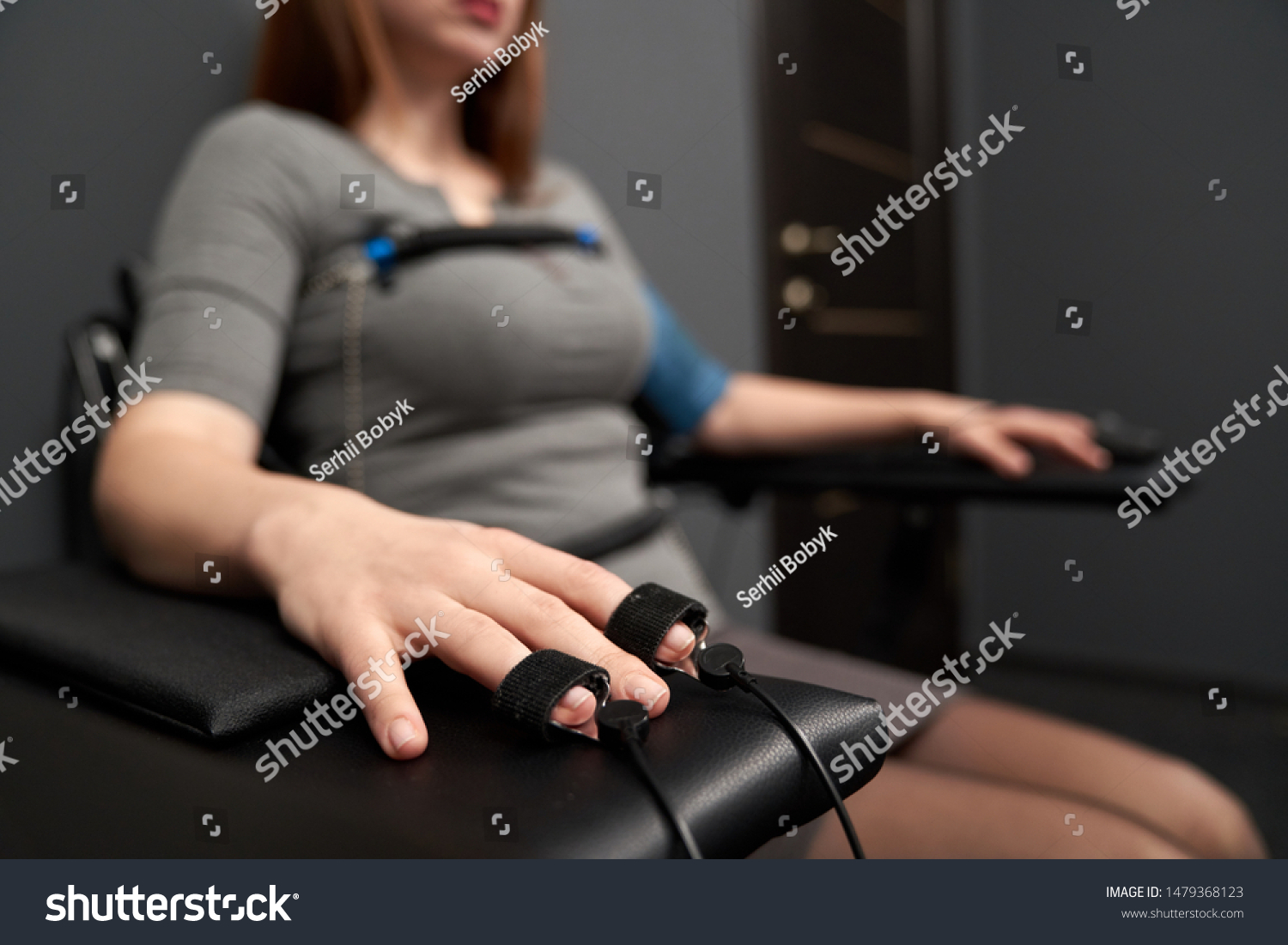Closeup of female hand with indicators measuring pulse during lie detector test. Sincere woman in grey dress sitting in chair and answering questions. Concept of examination and inquiry. #1479368123