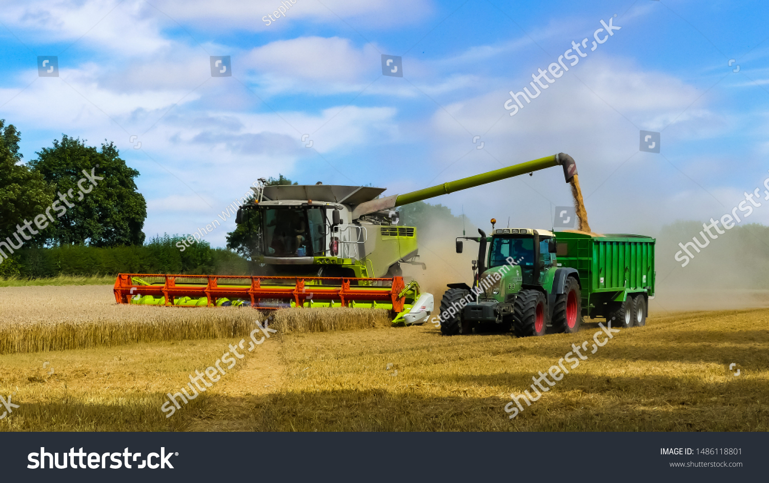 Tractor with trailer working in tandem alongside a working combine harvester discharging grain from uploader in an English cornfield. Dust clouds. Landscape image with space for text. Oxfordshire. #1486118801