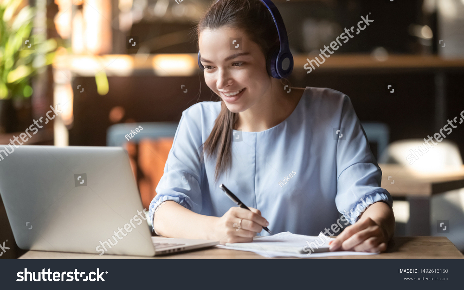 Smiling girl student wear wireless headphone study online with skype teacher, happy young woman learn language listen lecture watch webinar write notes look at laptop sit in cafe, distant education #1492613150