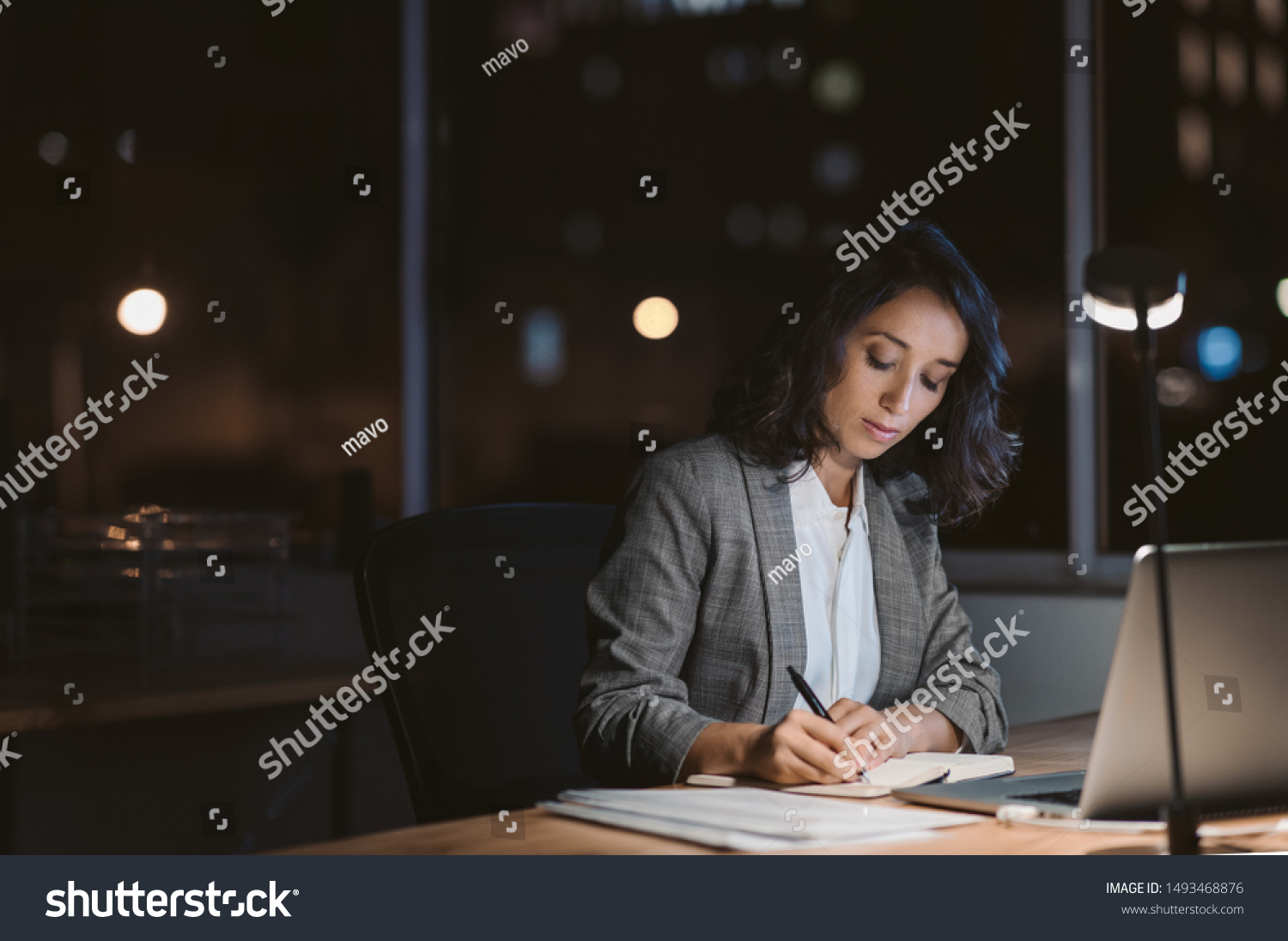 Young businesswoman using a laptop and writing notes while working overtime at her office desk late in the evening #1493468876