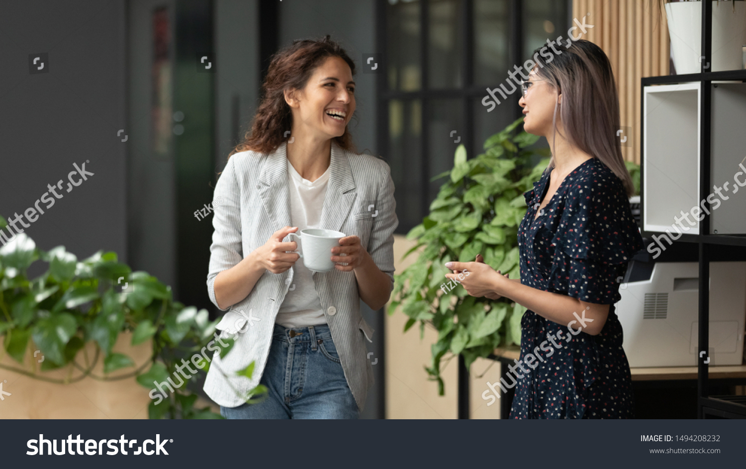 Happy positive female colleagues joking laughing during coffee break in work space, smiling diverse women business team talking having fun enjoy conversation good friendly relations walking in office #1494208232