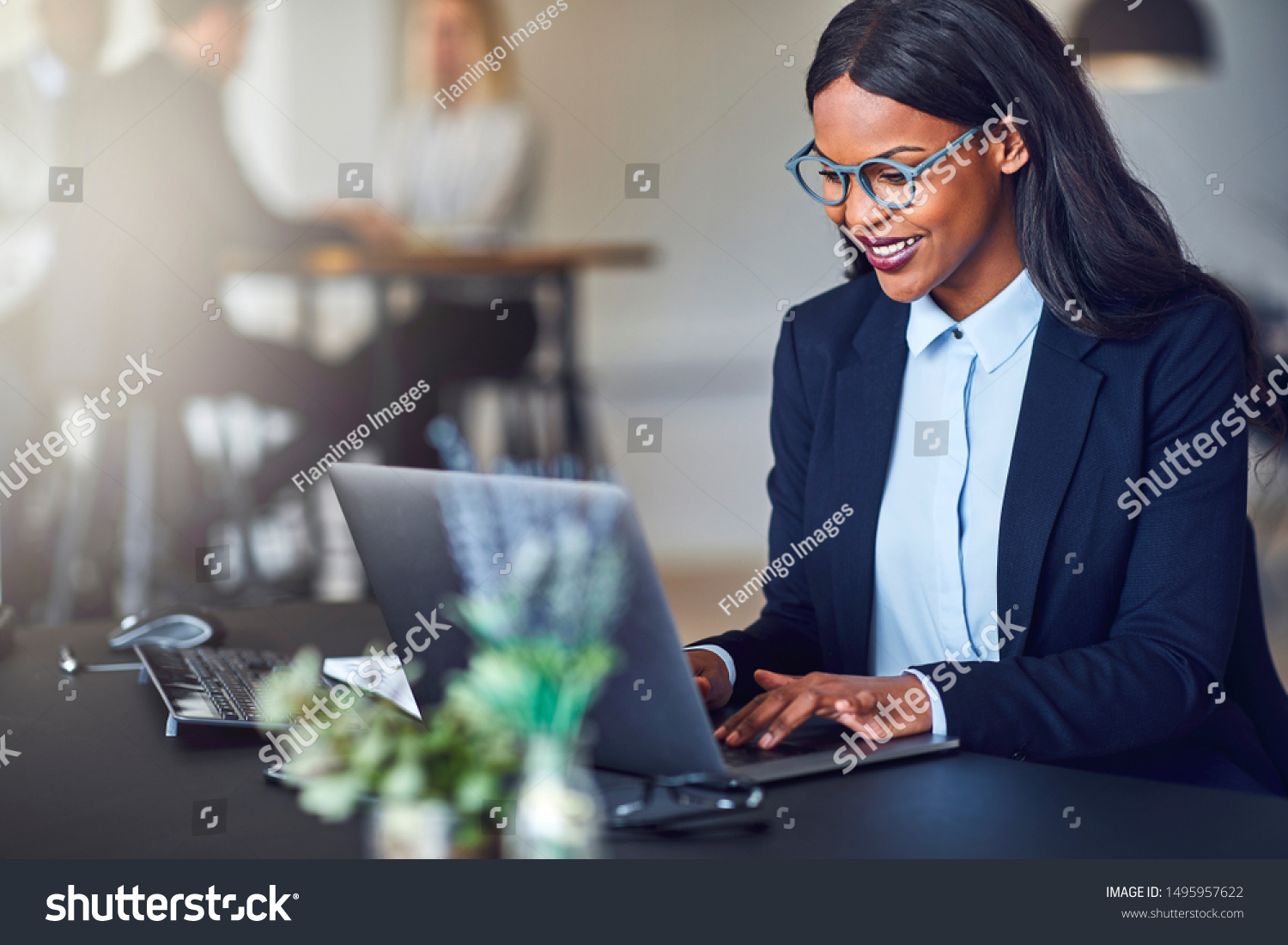 Smiling young African American businesswoman working on a laptop at her desk in a bright modern office with colleagues in the background #1495957622