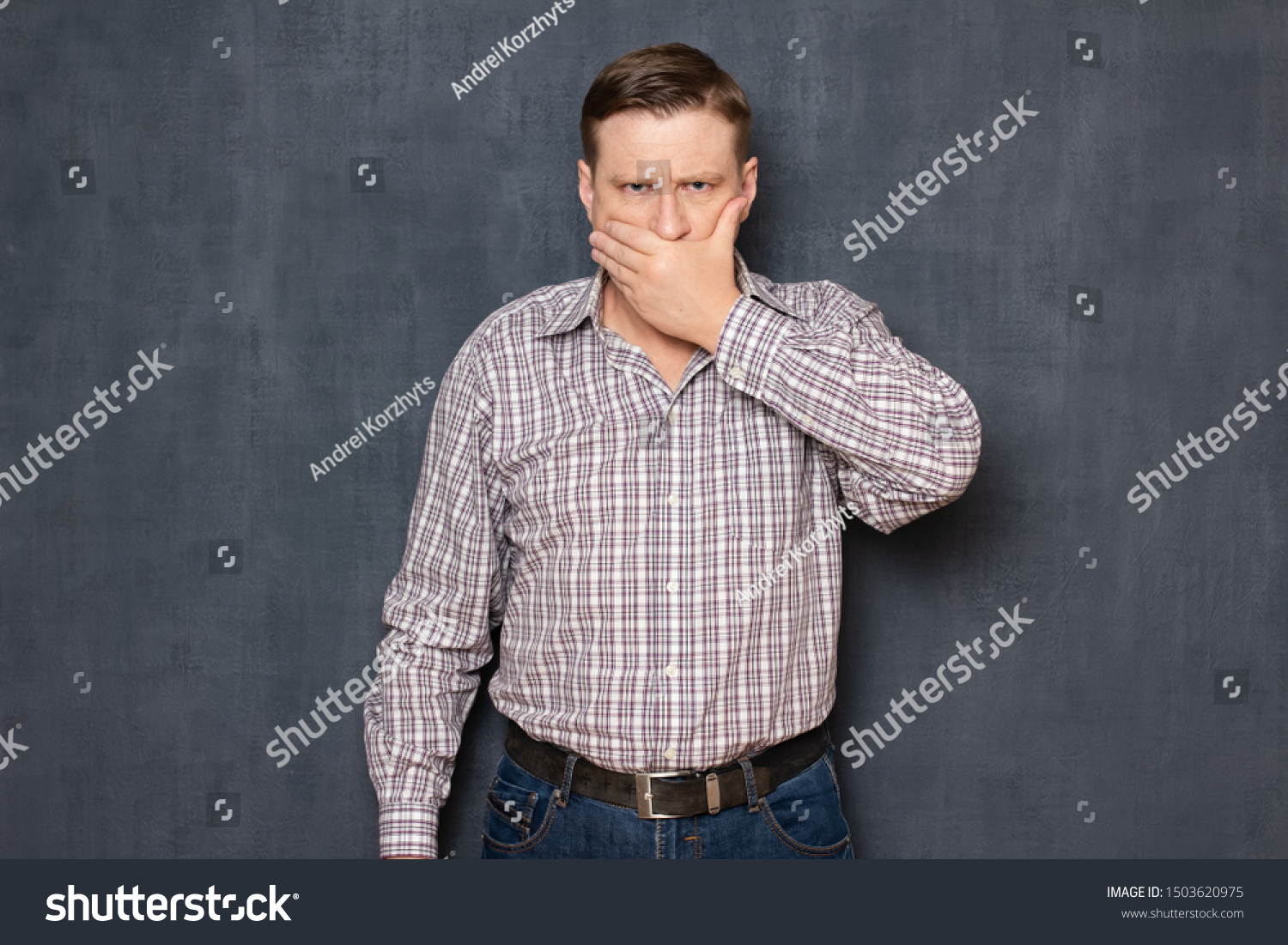 Studio half-length portrait of angry man wearing checkered shirt, covering mouth with hand like not wanting to say something, over gray background. Concept of deliberate silence and reticence #1503620975