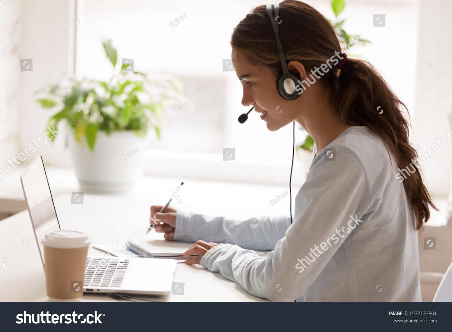 Side view concentrated millennial mixed race woman wearing headset with microphone, looking at laptop screen, listening educational webinar, taking notes, getting remote online education at home. #1537133861