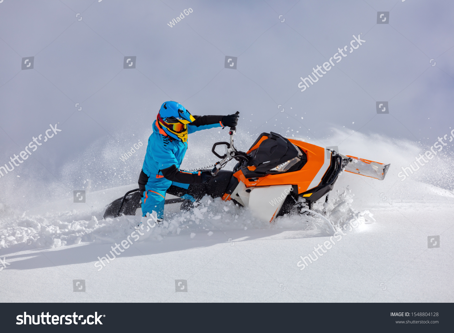 the guy turns a snowmobile in a mountain valley on the background of the clear snow and sky, leaving behind a trail of splashes. bright snow bike and suit without brands. Boondocker sports snowmobile #1548804128