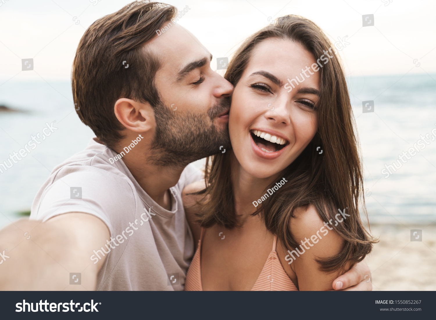 Image of young happy man kissing and hugging beautiful woman while taking selfie photo on sunny beach #1550852267