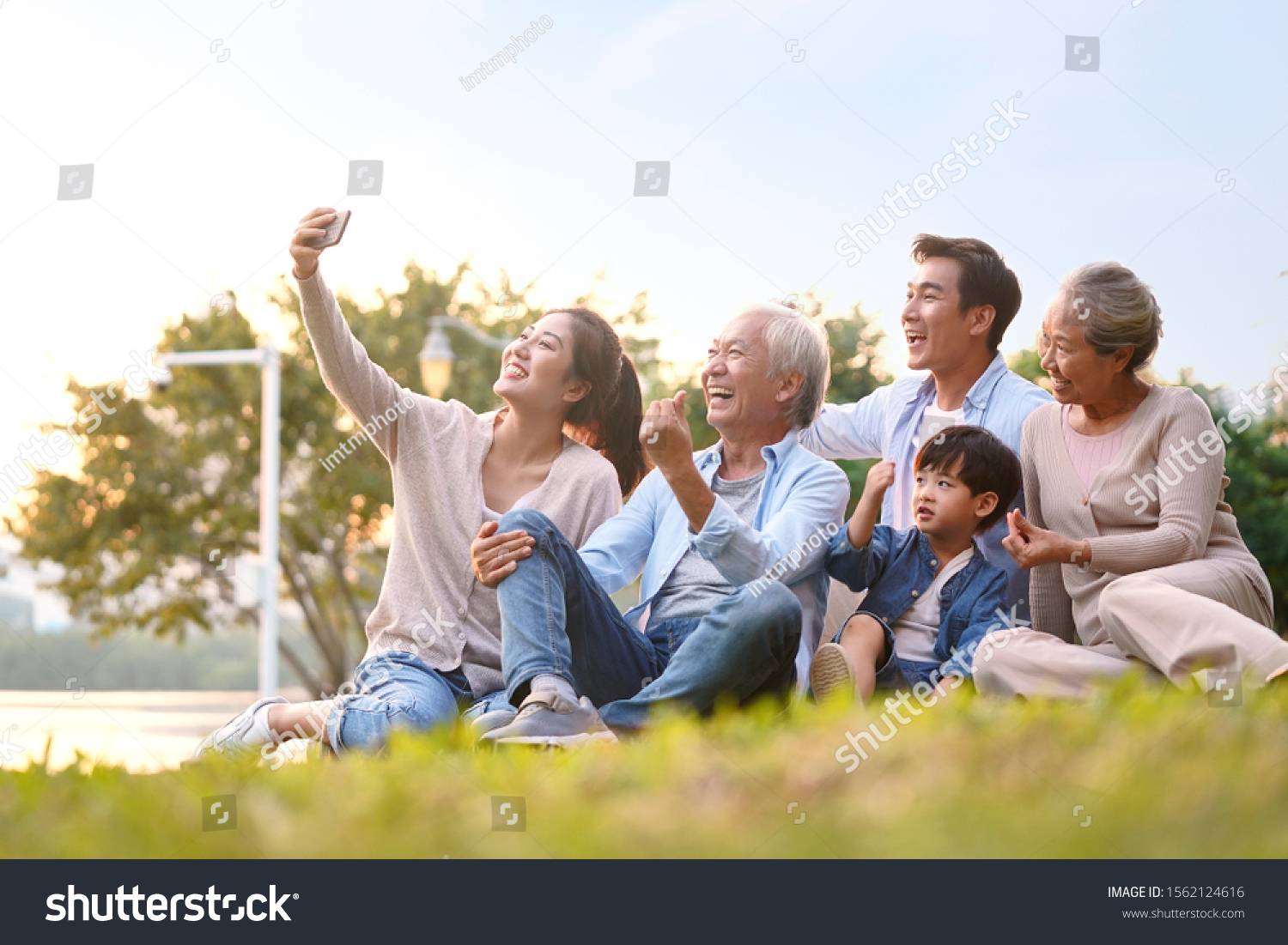 three generation happy asian family sitting on grass taking a selfie using mobile phone outdoors in park #1562124616