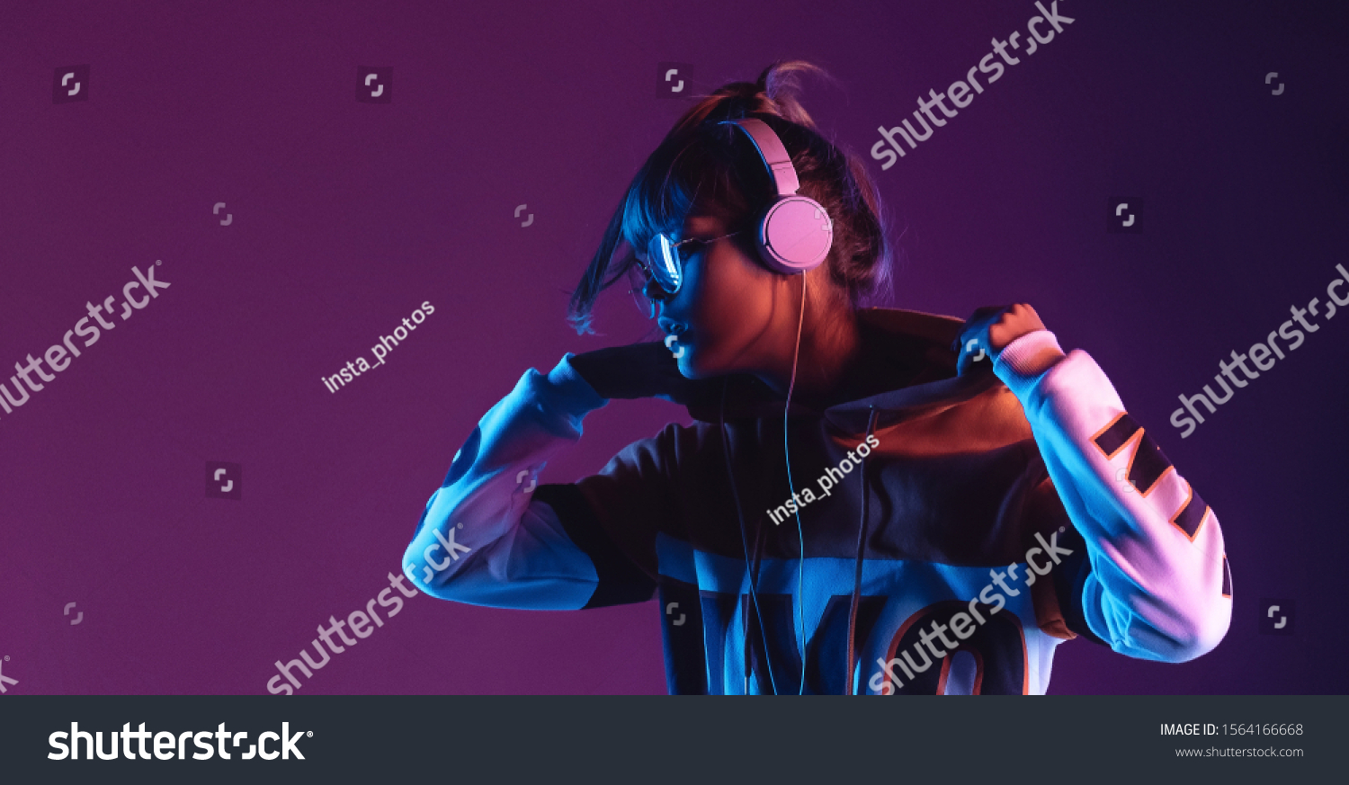 Hipster igen teen pretty fashion girl model wear stylish glasses headphones enjoy listen new cool music mix stand at purple studio background in trendy 80s 90s club blue party light, profile view #1564166668