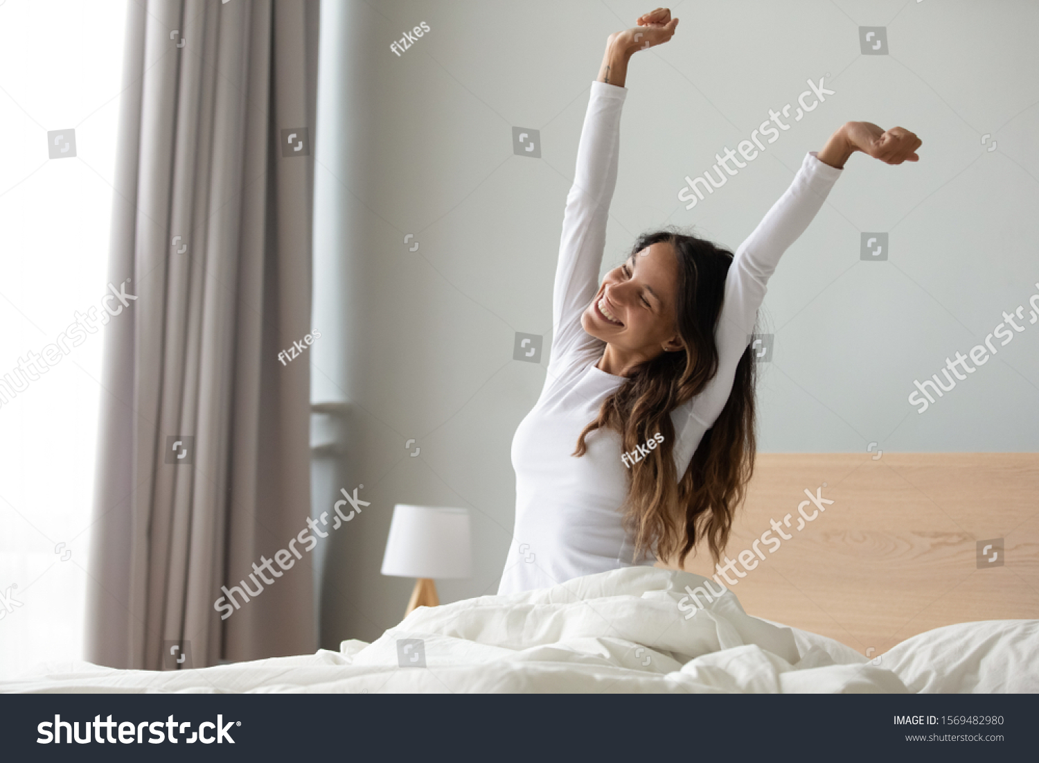 Happy woman in white nightwear sitting in bed awakened from enough and healthy sleep feels good, stretching her arms muscles after sleep and long immobility wakes up start new day with smile concept #1569482980