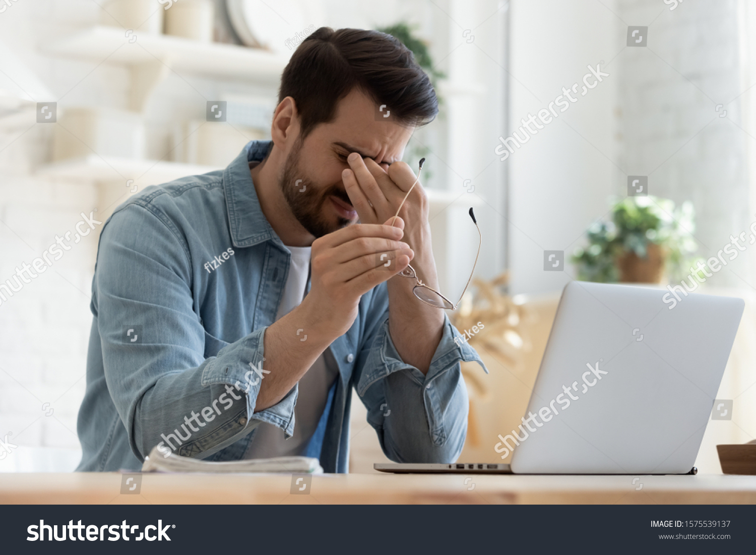 Tired young man feel pain eyestrain holding glasses rubbing dry irritated eyes fatigued from computer work, stressed man suffer from headache bad vision sight problem sit at home table using laptop #1575539137