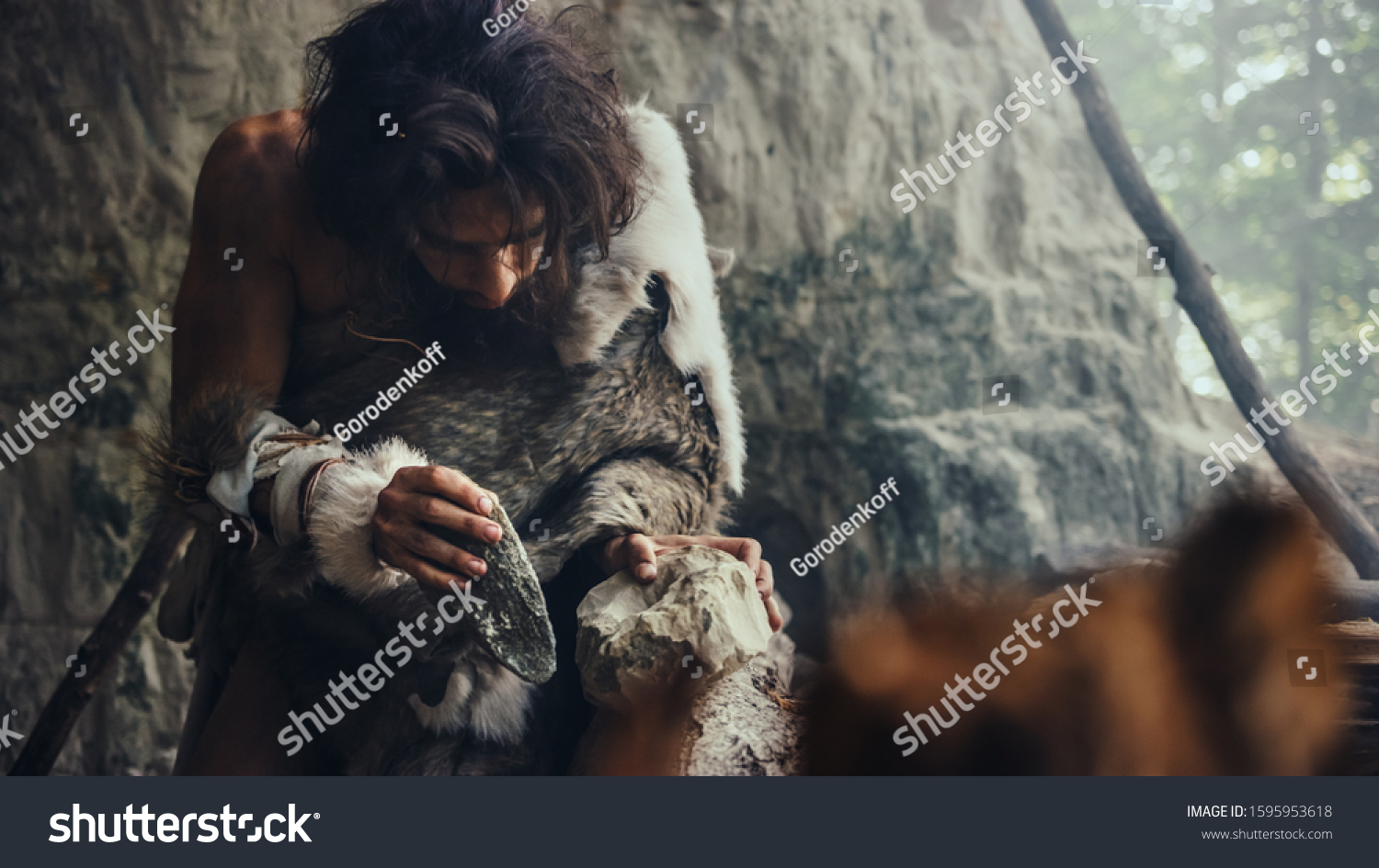 Close-up Shot of a Primeval Caveman Wearing Animal Skin Hits Rock with Sharp Stone, Makes First Primitive Tool for Hunting Animal Prey. Neanderthal Using Flint Rock. Dawn of Human Civilization. #1595953618
