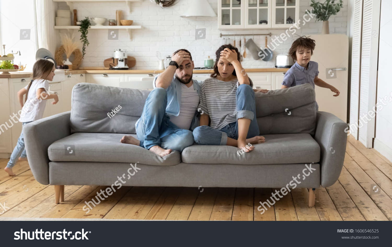 Tired mother and father sitting on couch feels annoyed exhausted while noisy little daughter and son shouting run around sofa where parents resting. Too active hyperactive kids, need repose concept #1606546525