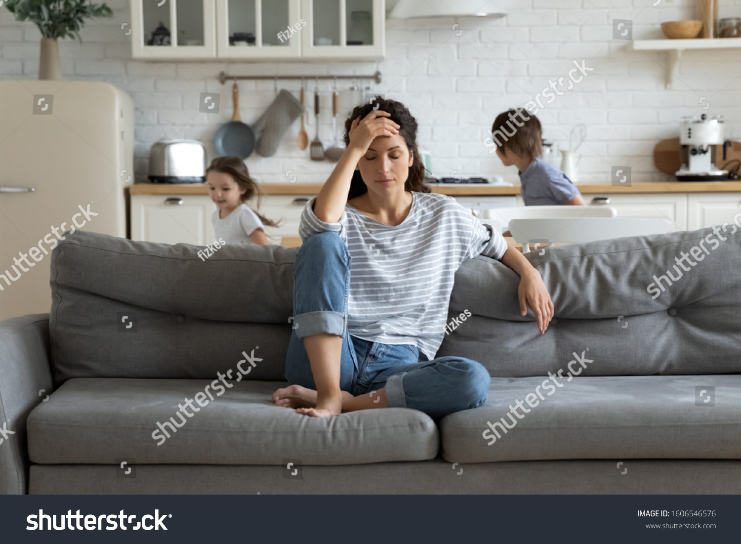 Young tired single mother suffers from headache closed eyes touch forehead sitting on couch while her daughter and son running around her and shouting, female babysitter feels exhausted by noisy kids #1606546576