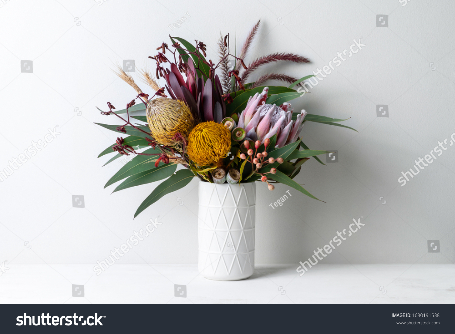 Beautiful floral arrangement of mostly Australian native flowers, including protea, banksia, kangaroo paw, eucalyptus leaves and gum nuts, in a white vase on a white table with a white background. #1630191538