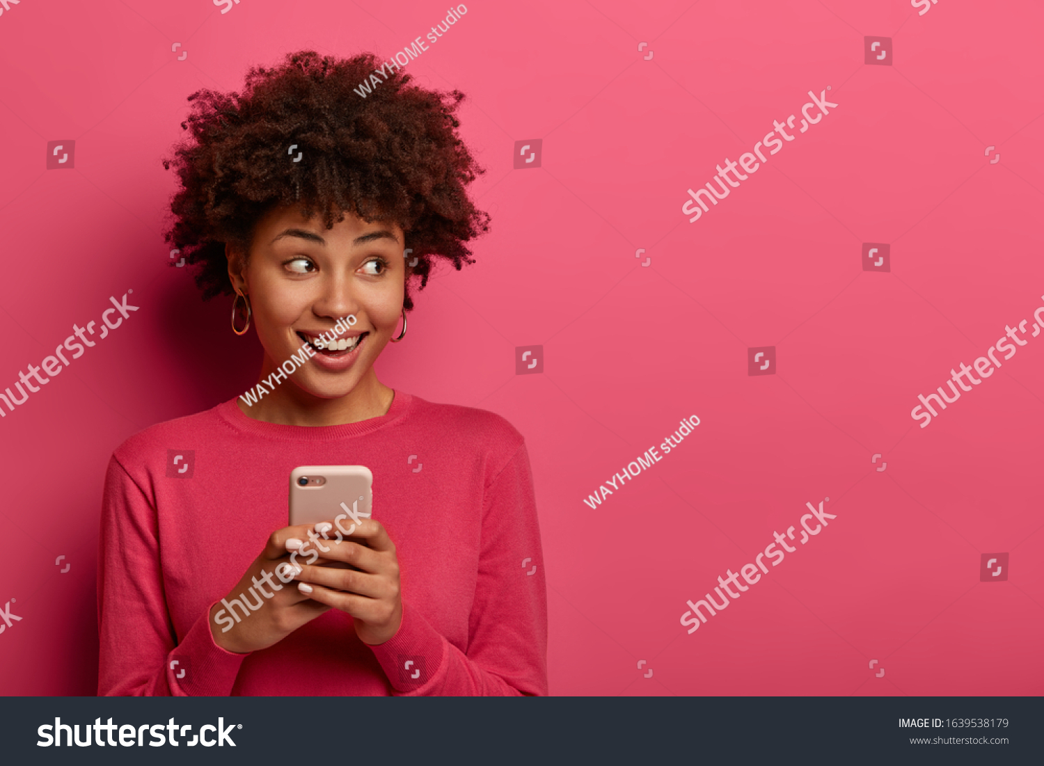 Portrait of cute curly haired girl scrolls social networks or does shopping online, looks aside positively, uses modern smartphone, wears pink sweater, poses indoor in studio, reads message. #1639538179
