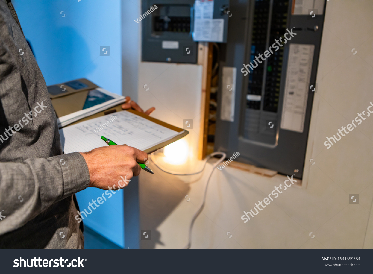 home Inspector in front of electric distribution board during inspection, selective focus and close up on man's hands as he holding notebook and pen #1641359554