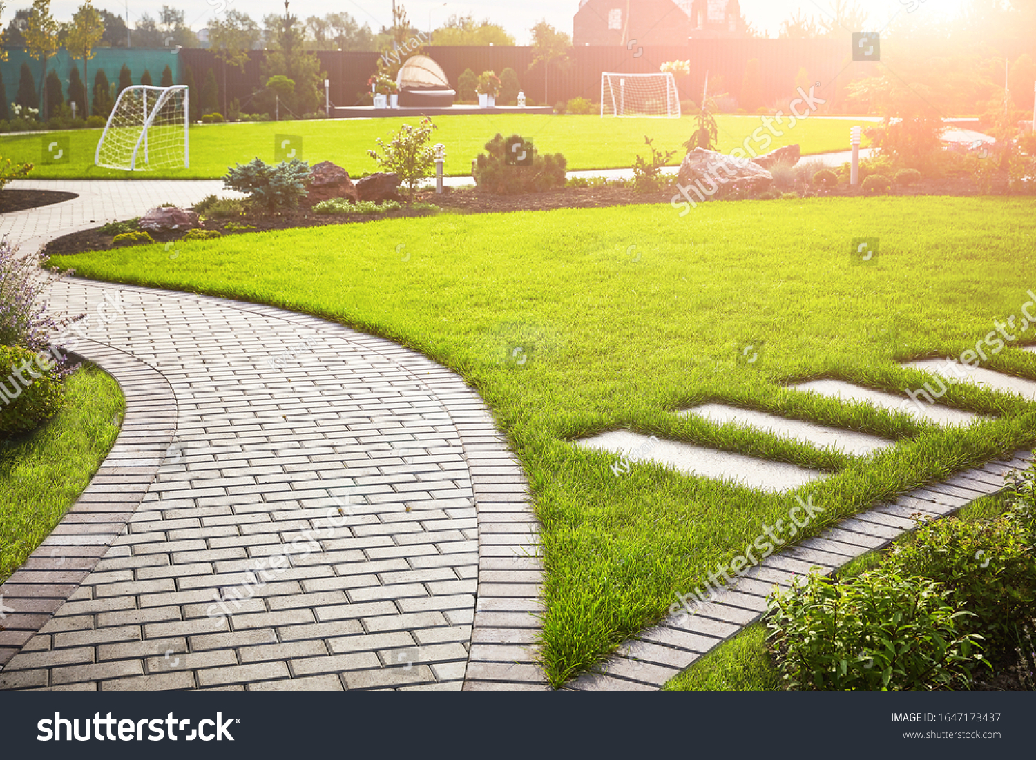 Landscaping of the garden. A tile path between green grass and a lawn with flowers in the sun. Soccer field in the background with copy space. #1647173437