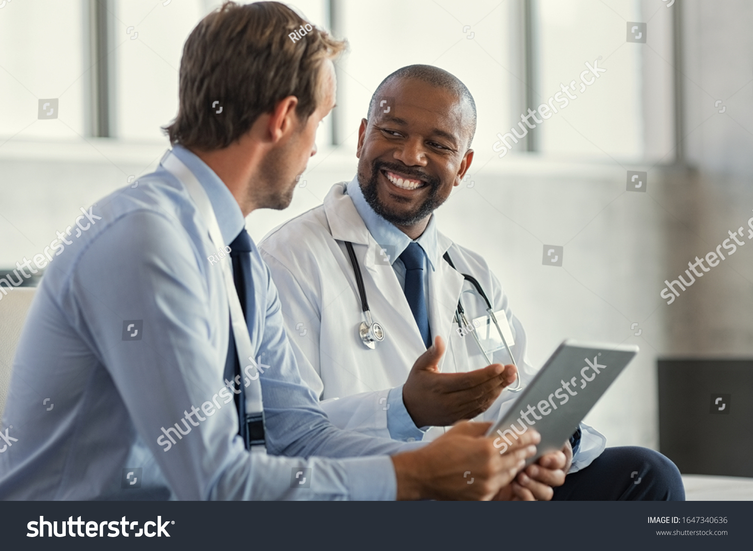 Two mature smiling doctors having discussion about patient diagnosis, holding digital tablet. Representative pharmaceutical discussing case after positive result with happy doctor about new medicine. #1647340636