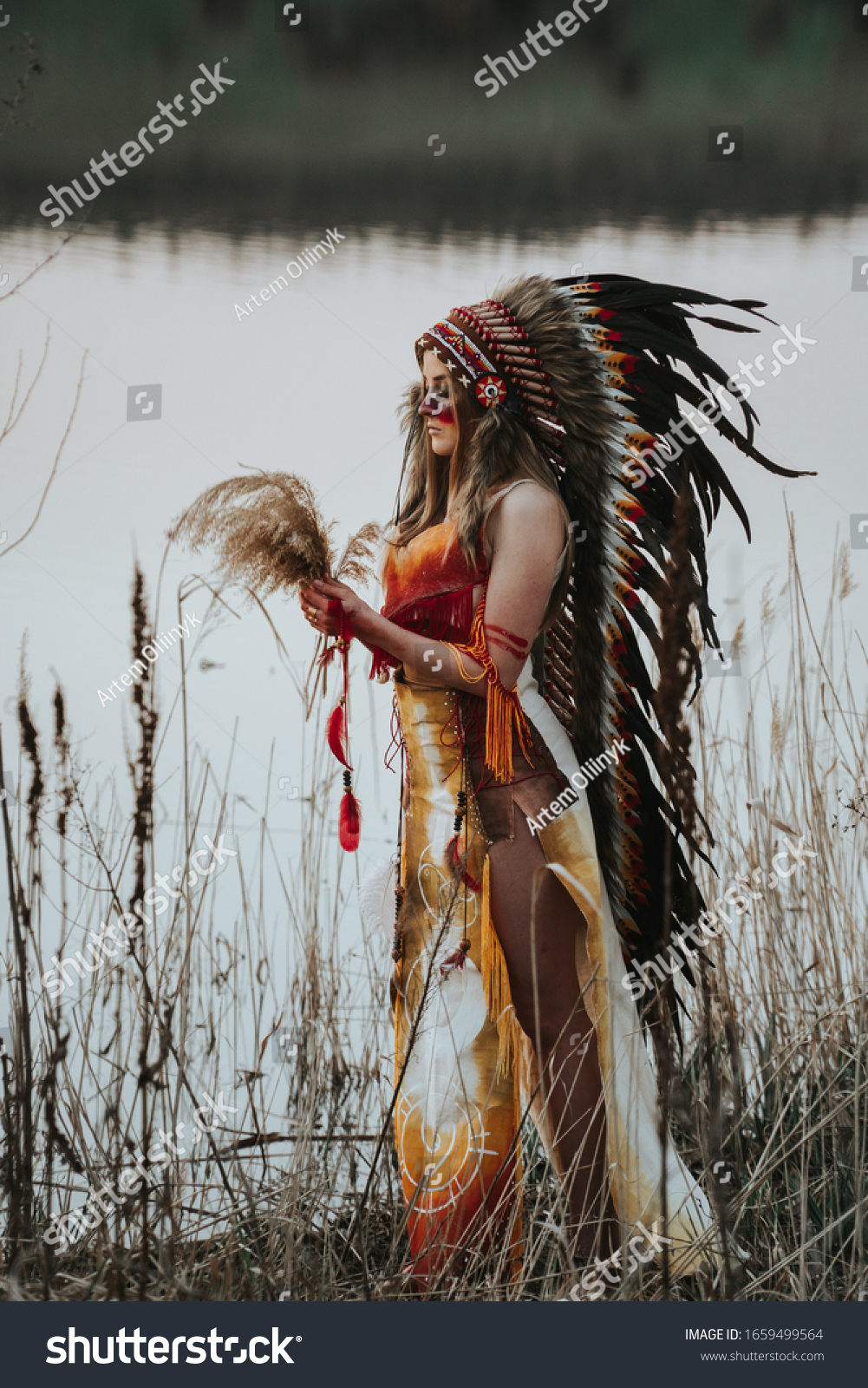 A girl dressed as a Native American with herbs in hands stands on the bank of the river #1659499564