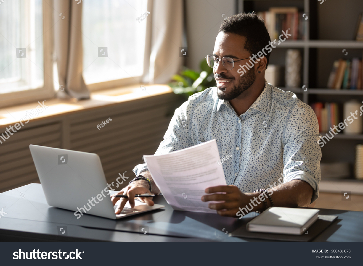Happy millennial african american businessman in eyewear holding documents, doing paperwork, preparing report or analyzing market research results, working on computer in modern workplace office. #1660489873
