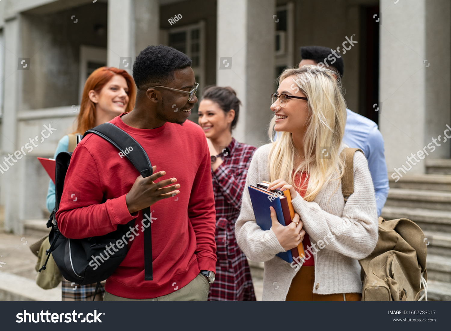 Group of happy multiethnic students walking outside college building. Five university students holding books and school backpack while talking together. Young man and beautiful girl in conversation. #1667783017