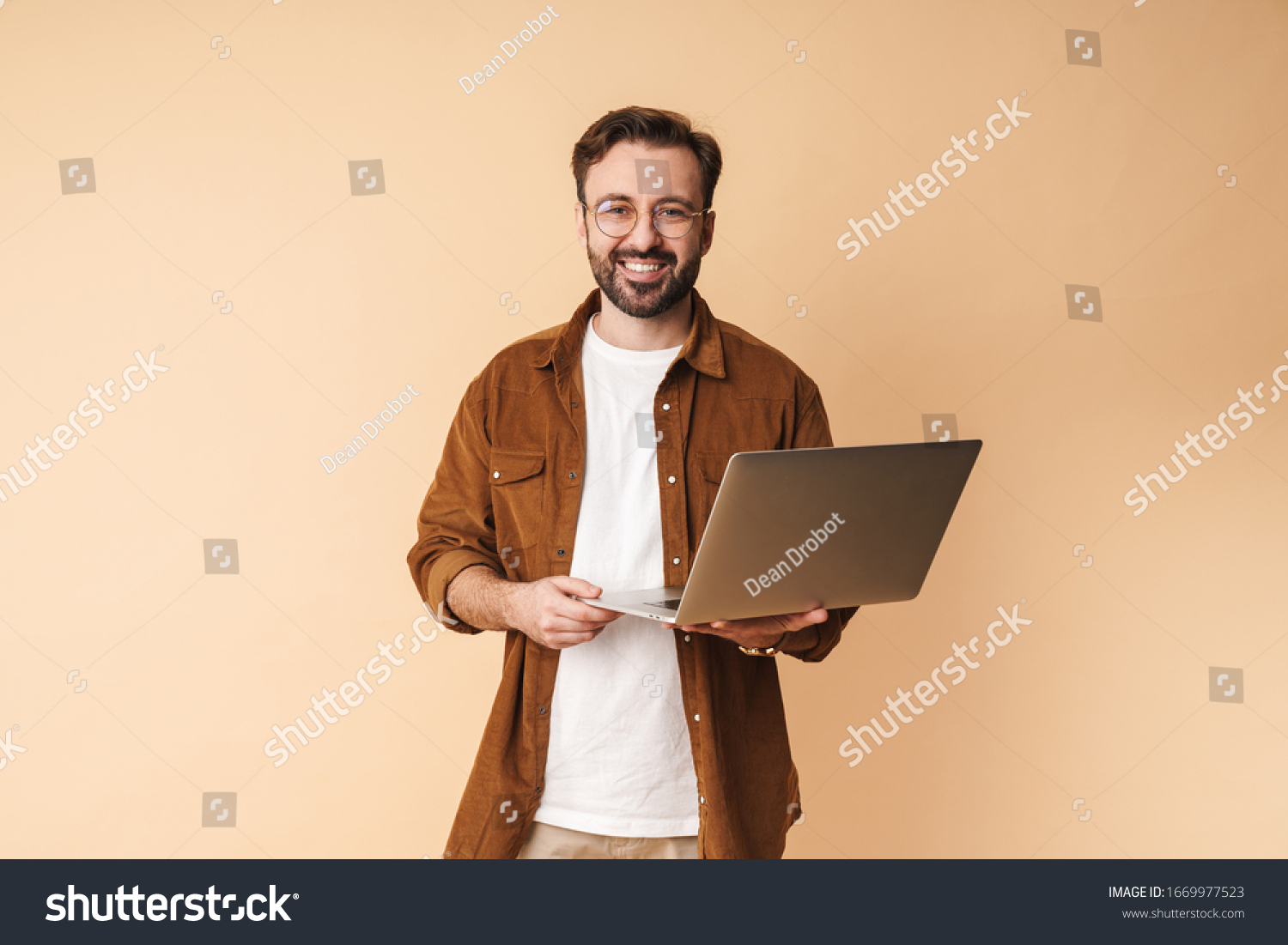 Image of a happy cheery optimistic young unshaved man isolated over beige wall background using laptop computer. #1669977523