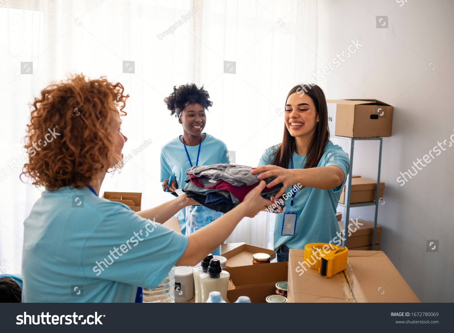 Volunteers Collecting Food Donations In Warehouse. Team of volunteers holding donations boxes in a large warehouse. Volunteers putting clothes in donation boxes, social worker making notes charity #1672780069