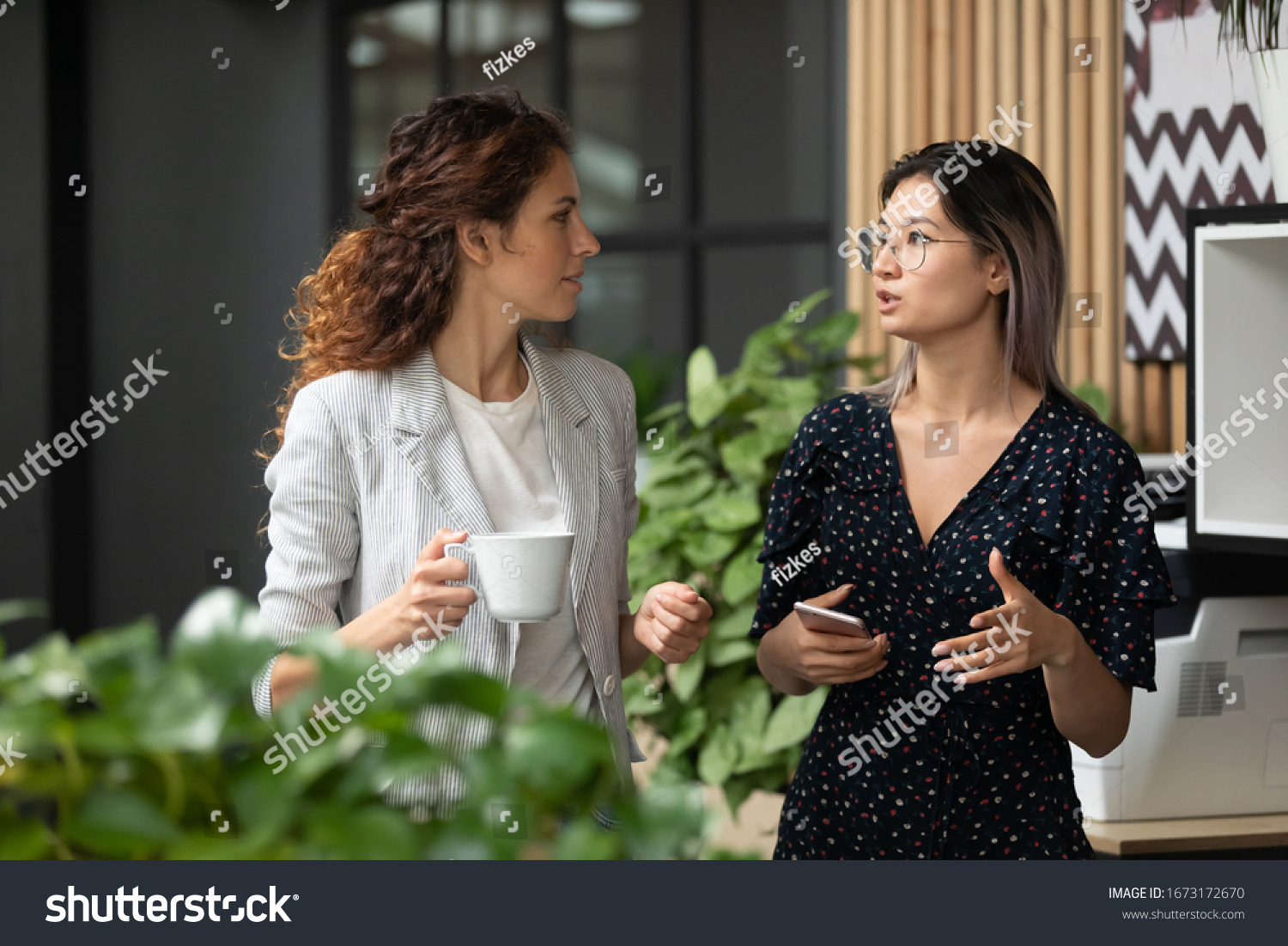 Diverse employees chatting during coffee break, walking in modern office, Asian businesswoman wearing glasses sharing ideas, discussing project with colleague, having pleasant conversation #1673172670