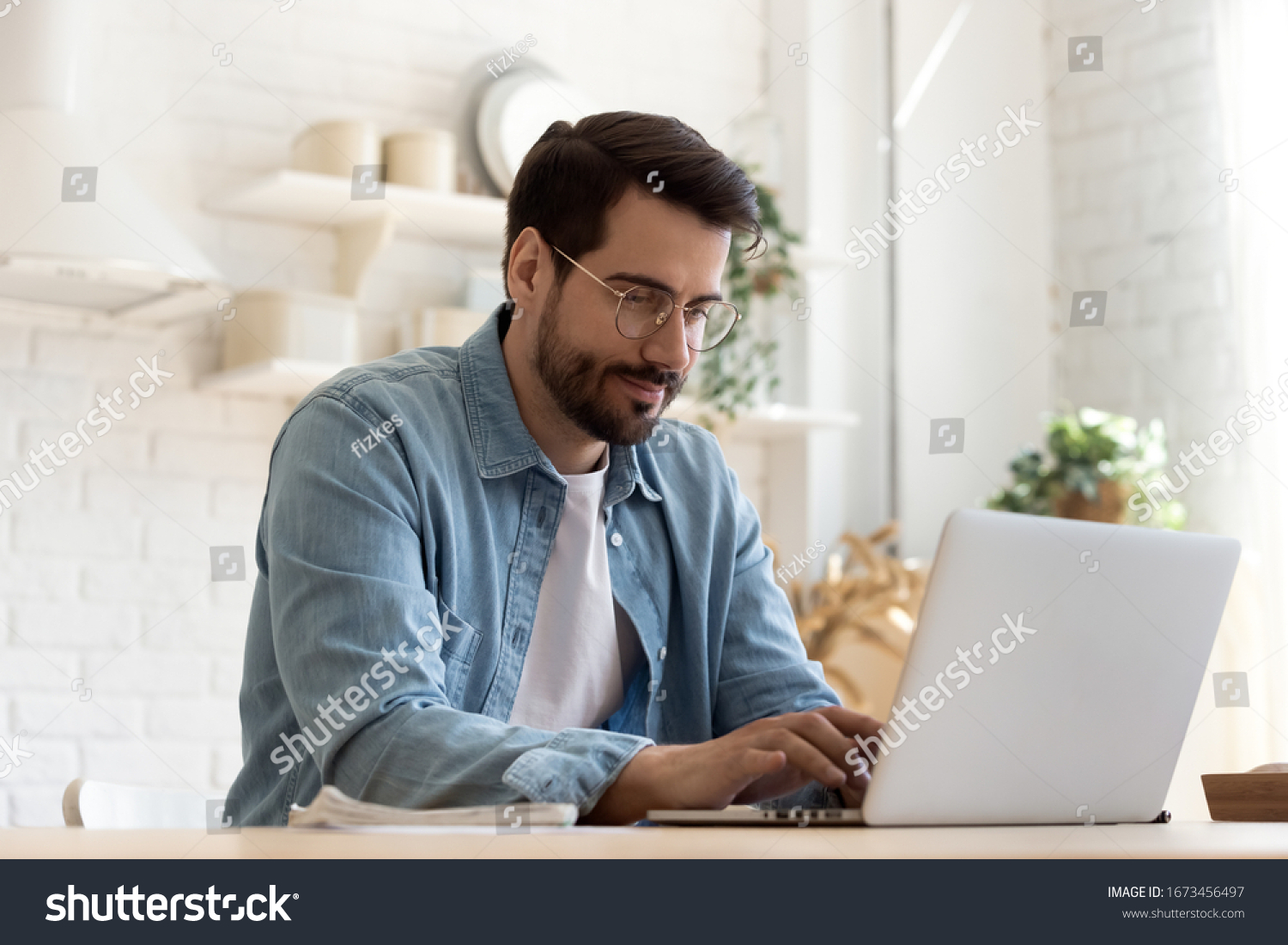 Focused young man wearing glasses using laptop, typing on keyboard, writing email or message, chatting, shopping, successful freelancer working online on computer, sitting in modern kitchen #1673456497