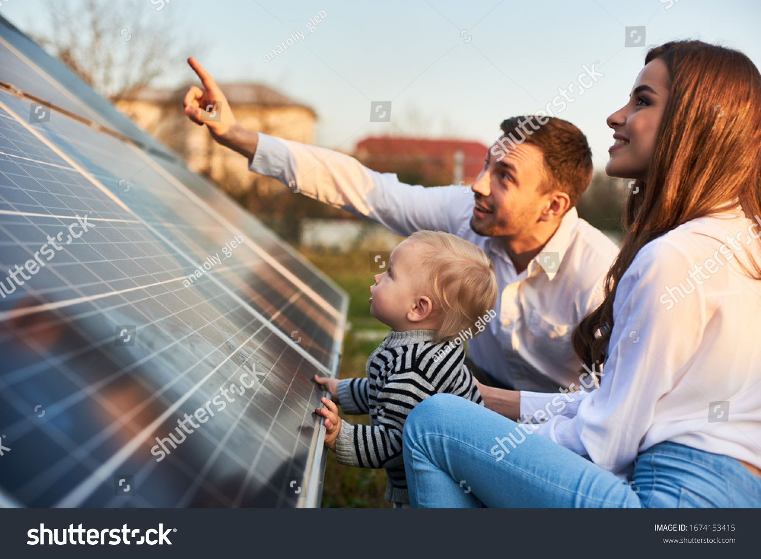 Man shows his family the solar panels on the plot near the house during a warm day. Young woman with a kid and a man in the sun rays look at the solar panels. #1674153415