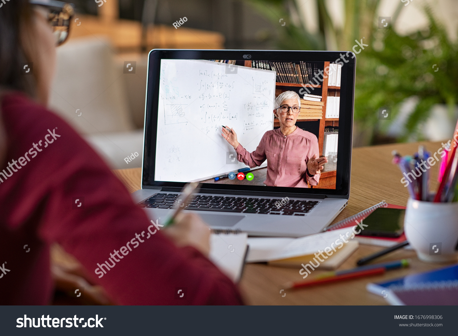 Young student watching lesson online and studying from home. Young woman taking notes while looking at computer screen following professor doing math on video call. Girl studying from home on pc. #1676998306