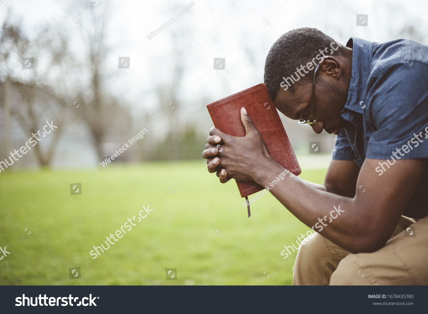A young African-American male sitting with closed eyes with the Bible in his hands #1678435780