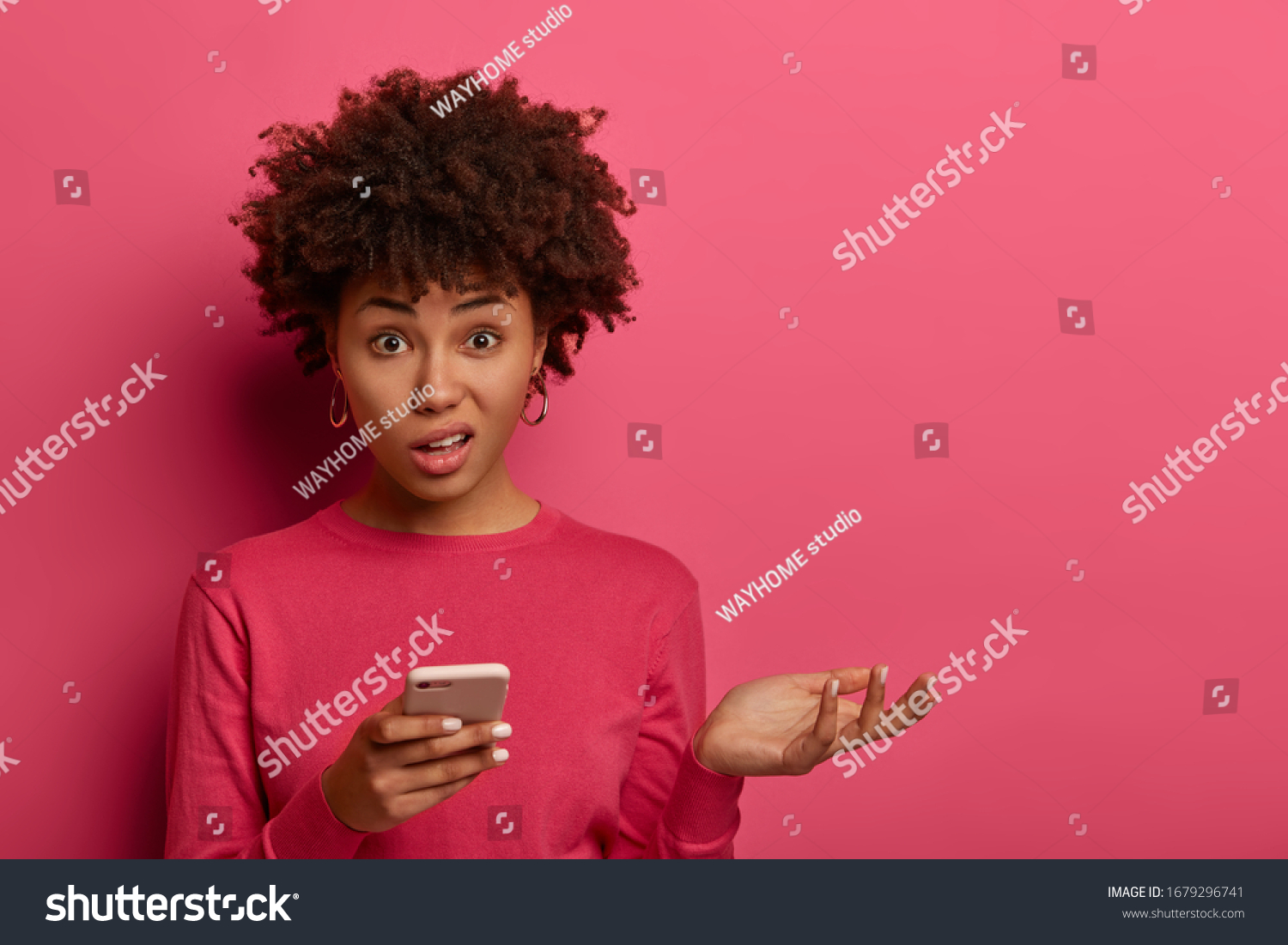 Distrubed dark skinned woman has problematic concerned face expression, holds smartphone, raises palm, cannot understand whats wrong, strange meaning of message, shrugs shoulders, stands indoor #1679296741