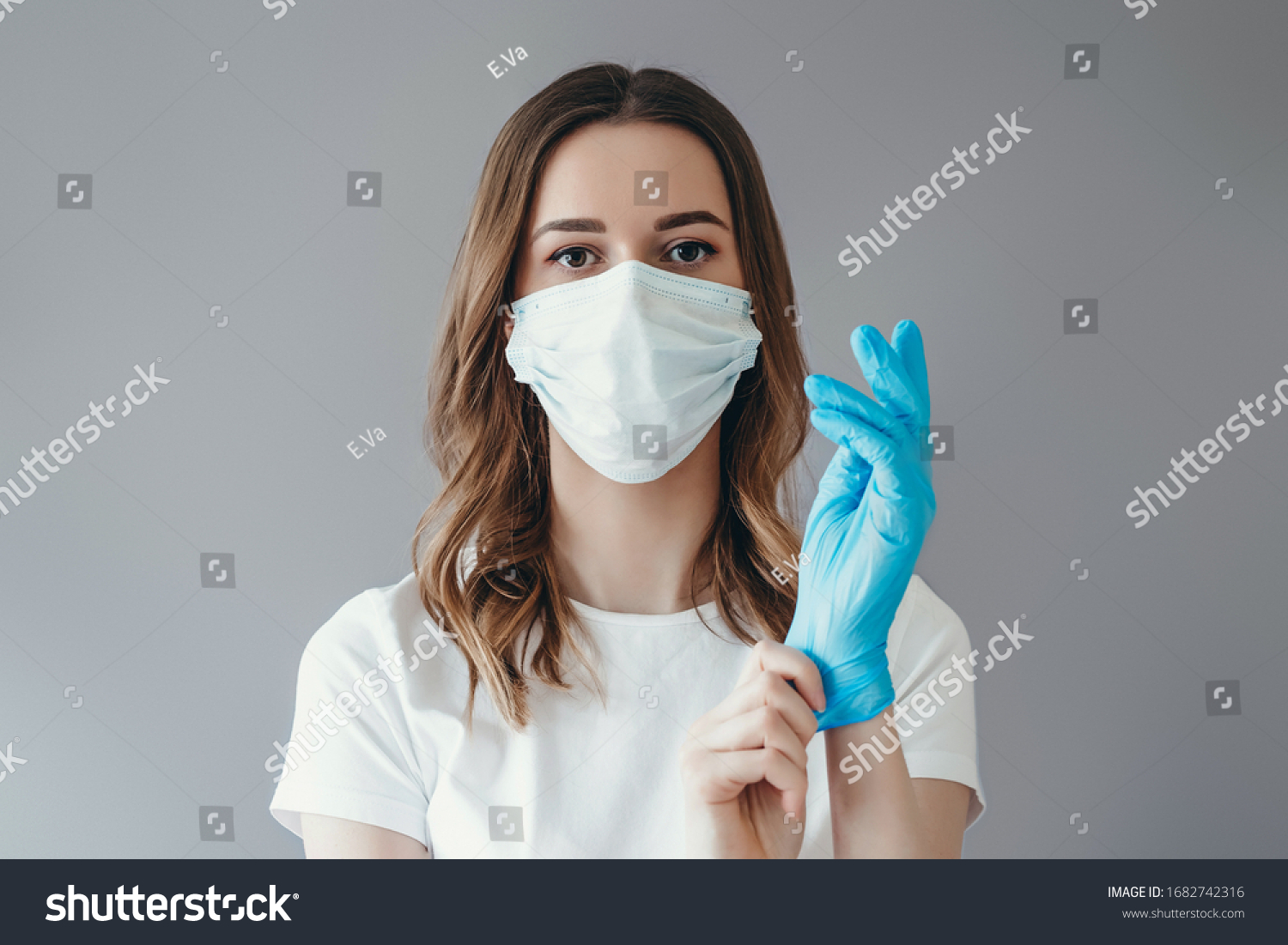 Young woman patient in a medical mask puts on protective surgical sterile gloves on her arm, isolated on gray background, protection against coronovirus #1682742316