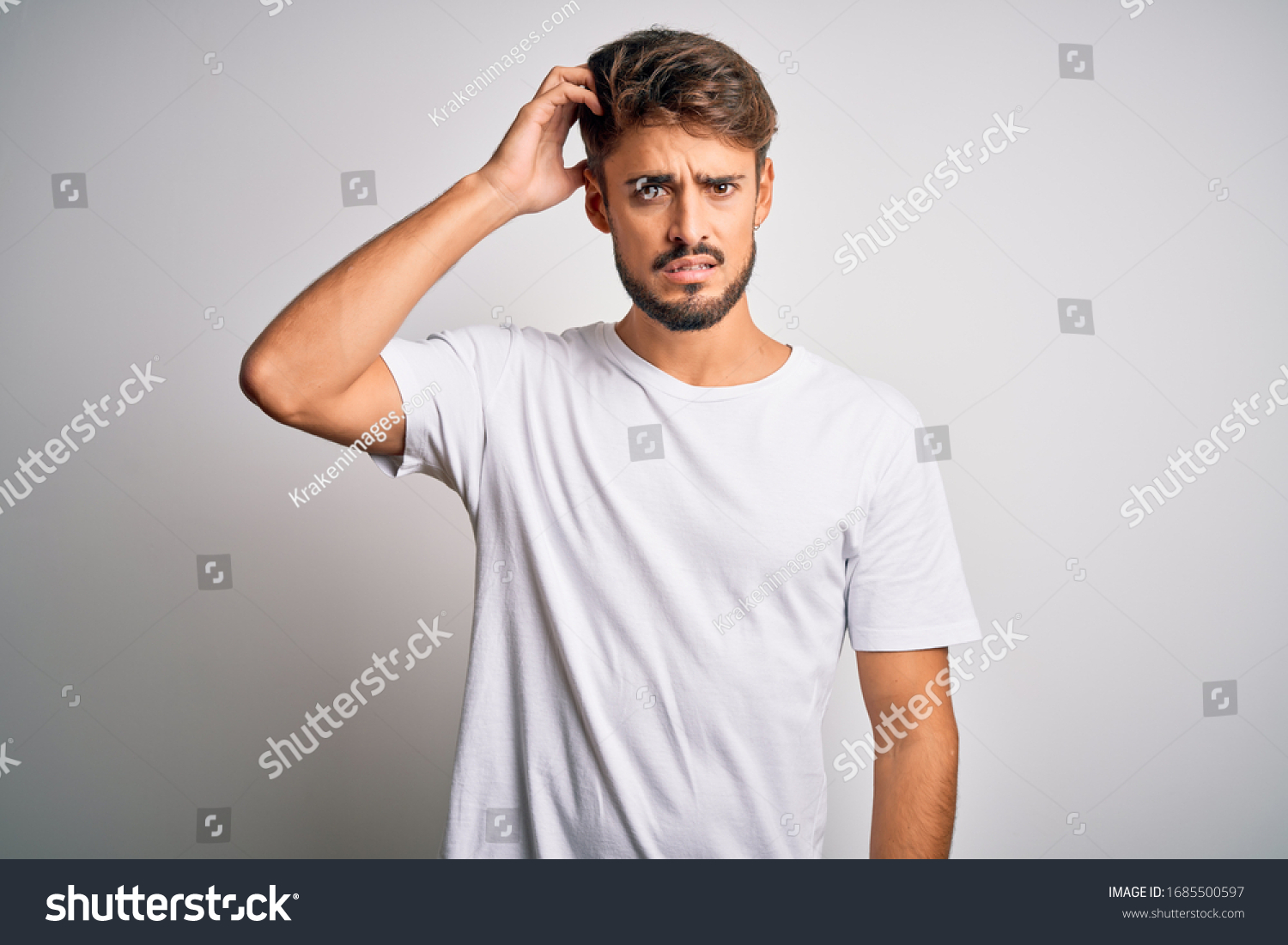 Young handsome man with beard wearing casual t-shirt standing over white background confuse and wonder about question. Uncertain with doubt, thinking with hand on head. Pensive concept. #1685500597
