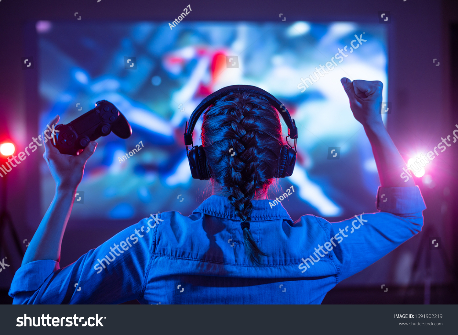 Girl in headphones plays a video game on the big TV screen. Gamer with a joystick. Online gaming with friends, win, prize. Fun entertainment. Teens play adventure games. Back view. Neon lighting #1691902219
