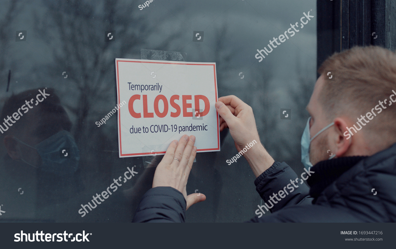 Caucasian male wearing medical mask puts a Temporary closed due COVID-19 pandemic sign on a window. Coronavirus pandemic, small business shutdown #1693447216