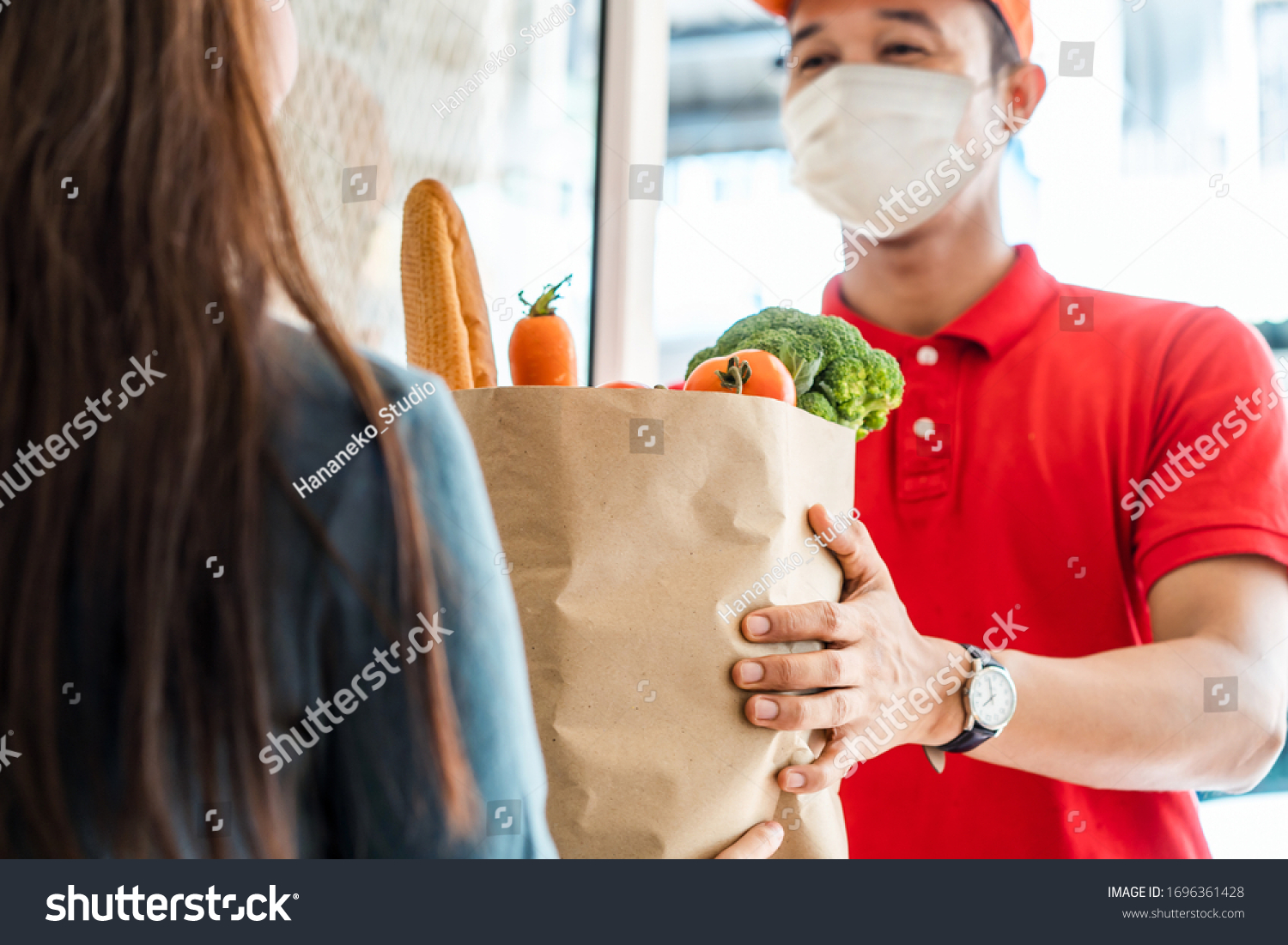Asian deliver man wearing face mask in red uniform handling bag of food, fruit, vegetable give to female costumer in front of the house. Postman and express grocery delivery service during covid19. #1696361428