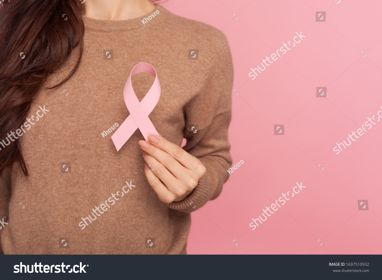 Female health check awareness. Closeup of young woman in pullover holding pink ribbon, symbol of breast cancer, timely diagnosis, support of oncology patients. studio shot isolated on pink background #1697510932