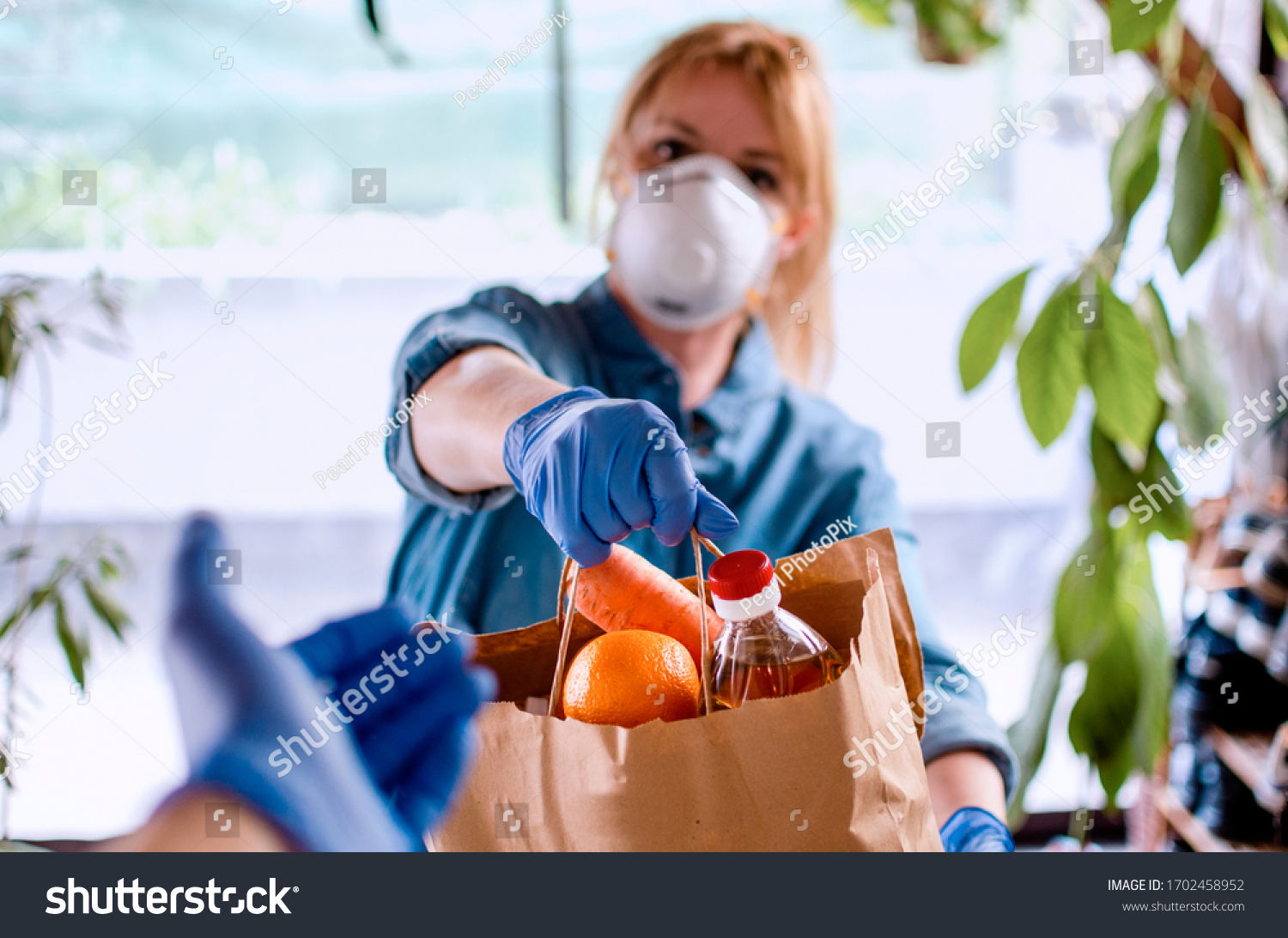 Woman delivering Food in paper bag during Covid 19 outbreak. Female  Volunteer holding groceries in the house porch. Delivery woman in face mask and gloves giving Donating neccessities to a hand #1702458952