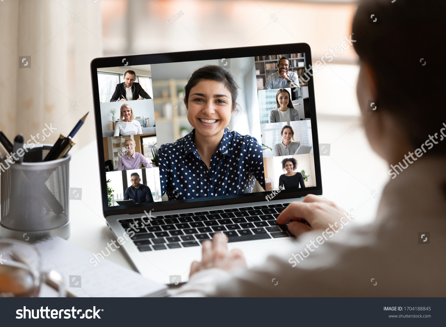Woman sit at desk looking at computer screen where collage of diverse people webcam view. Indian ethnicity young woman lead video call distant chat, group of different mates using videoconference app #1704188845