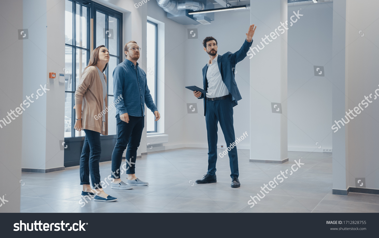 Real Estate Agent Showing a New Empty Office Space to Young Male and Female Hipsters. Entrepreneurs Meet the Broker with a Tablet and Discuss the Facility They Wish to Purchase or Rent. #1712828755