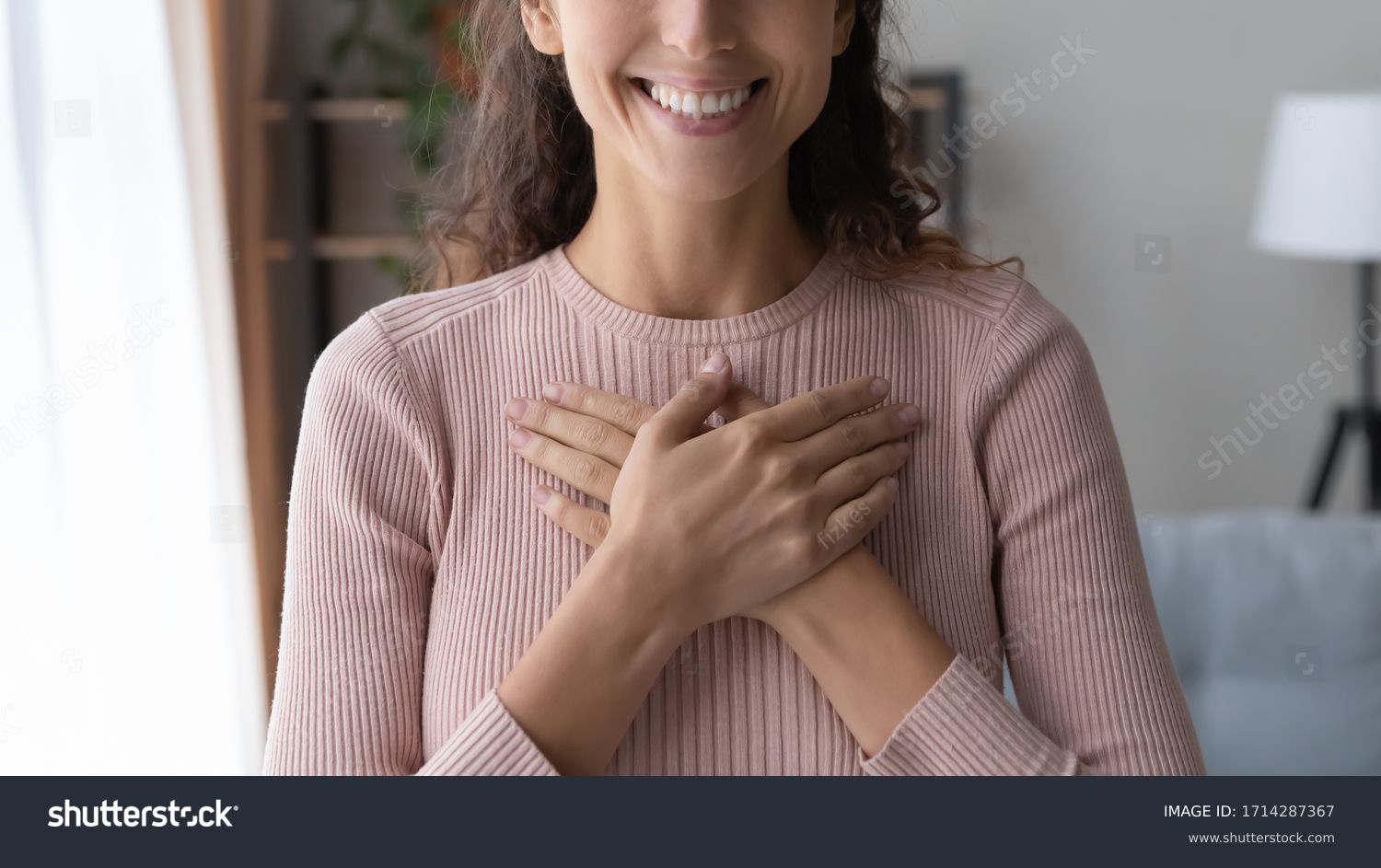 Close up focus on happy sincere female holding folded hands on chest. Emotional positive kind candid millennial woman feeling thankful indoors, showing gratitude sign, believe faith charity concept. #1714287367