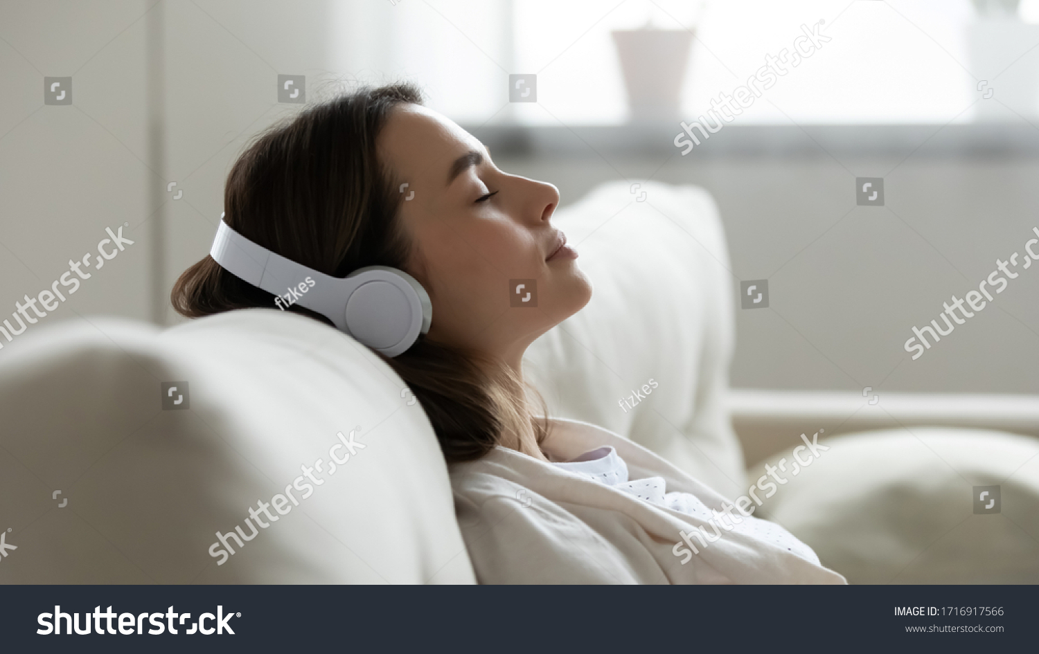 Peaceful girl in modern wireless headphones sit relax on comfortable couch listening to music, happy calm young woman in earphones rest on cozy sofa, enjoy good quality sound, stress free concept #1716917566
