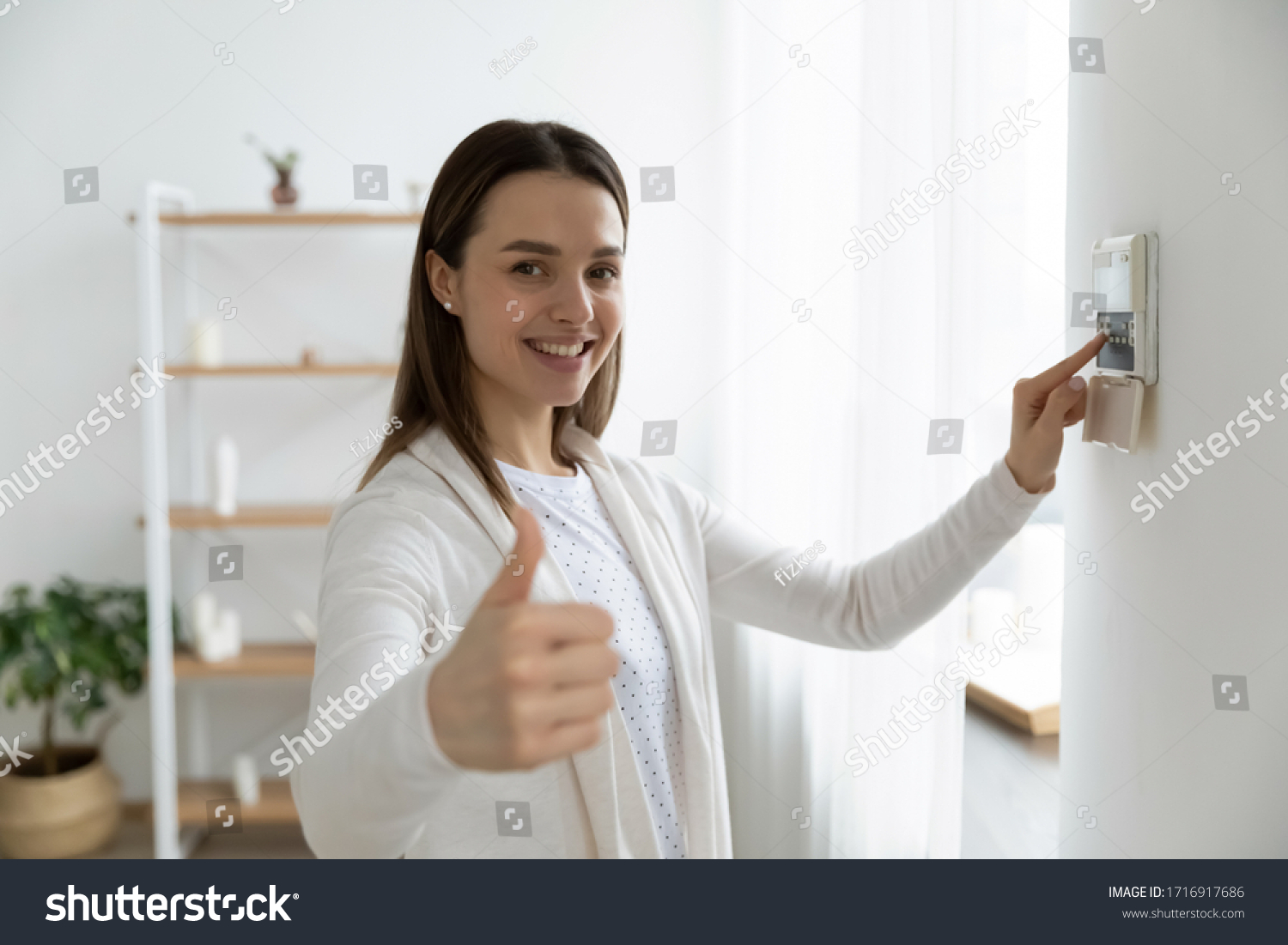 Smiling young woman use turn on smart home control panel on apartment wall, recommend good quality service show thumb up, happy millennial female client switch fire alarm or house security system #1716917686