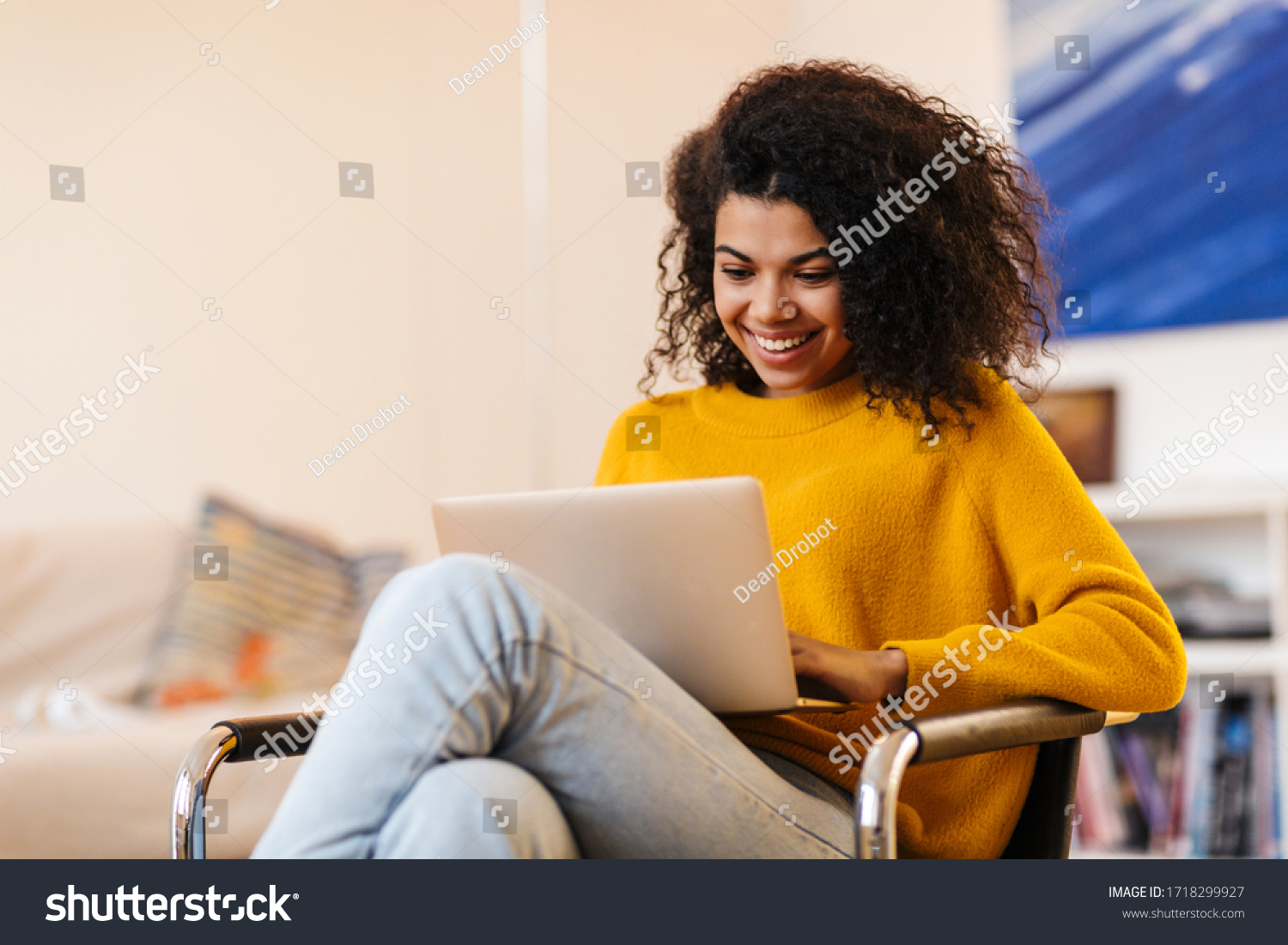 Image of cheerful african american woman using laptop while sitting on chair in living room #1718299927