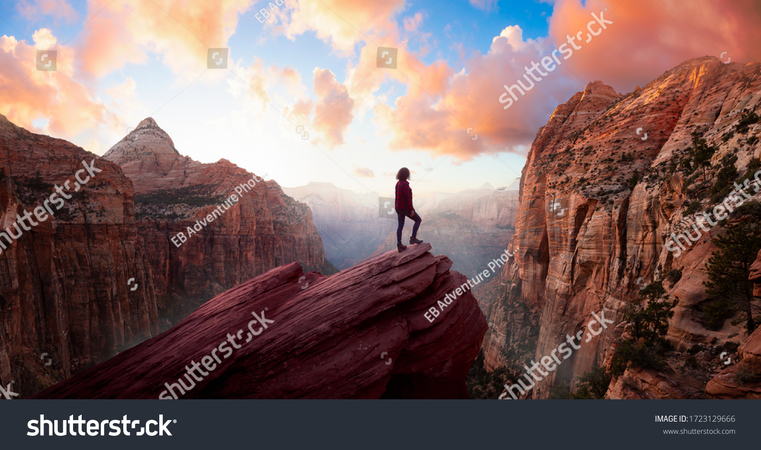 Adventurous Woman at the edge of a cliff is looking at a beautiful landscape view in the Canyon during a vibrant sunset. Taken in Zion National Park, Utah, United States. Sky Composite Panorama #1723129666