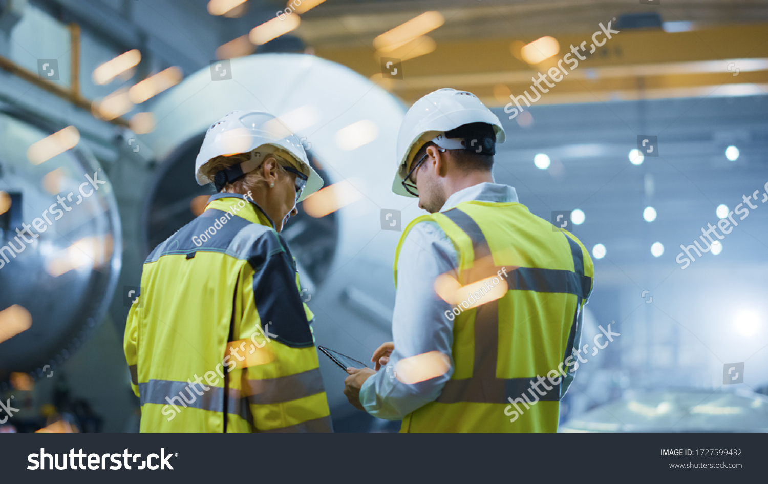 Two Heavy Industry Engineers Stand in Pipe Manufacturing Factory, Use Digital Tablet Computer, Have Discussion. Construction of Oil, Gas and Fuels Transport Pipeline. Back View Sparks Flying #1727599432